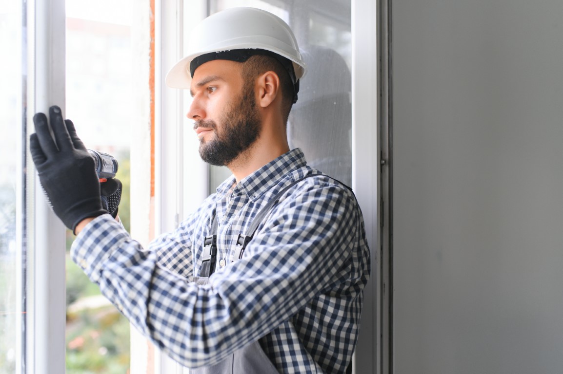Workman in overalls installing or adjusting plastic windows in the living room at home.